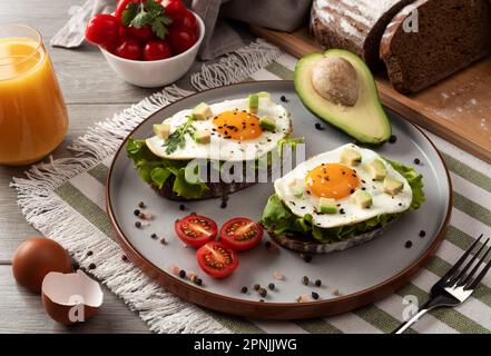 Toasts with avocado and fried eggs on table. Healthy breakfast with vegetables and herbs. Fried egg on a grey plate with orange juice. Serving on the Stock Photo