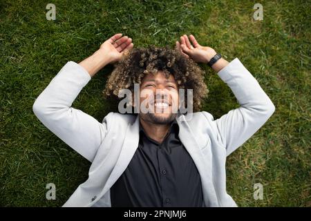 Happy black middle aged businessman in suit lying on green grass on lawn and relaxing in park, taking break from work Stock Photo