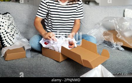 Unrecognizable woman packing a parcel box while sitting on sofa at home. Packing and unpacking, delivery service concept. Stock Photo