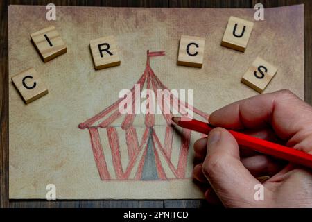 A male hand is coloring a drawing representing a circus tent with a red pencil Stock Photo