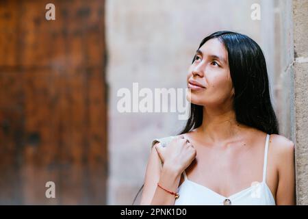 Dreamy Latin American woman adjusting backpack on shoulder and admiring street while leaning on wall of old building on street of Barcelona, Spain Stock Photo