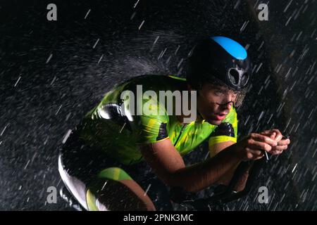 A triathlete braving the rain as he cycles through the night, preparing himself for the upcoming marathon. The blurred raindrops in the foreground and Stock Photo