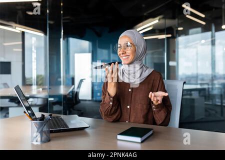 Young Muslim woman freelancer in hijab sitting at office desk in front of laptop and talking on speaker phone, recording conversation on phone. Stock Photo