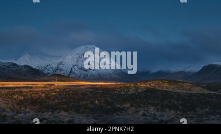 Night view of a road leading through the Scottish Highlands of Glen Coe with car light trail, snowcapped mountains and valley. Scotland Stock Photo