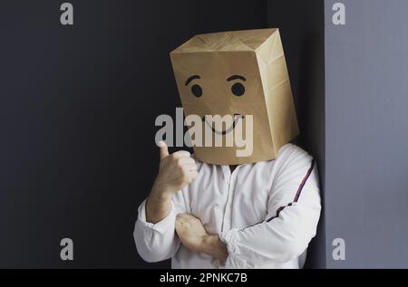 A man in a white shirt with a paper bag on his head, with a smiley face drawn, stands against the wall, overwhelmed with feelings, shows a hand gestur Stock Photo