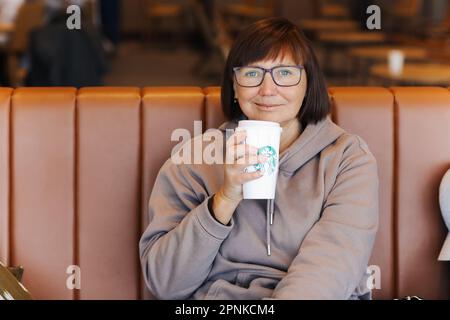 Valencia, Spain : March 18, 2023: Smiling middle aged woman in casual clothes holds a paper cup of hot Starbucks coffee with straw at Starbucks coffee Stock Photo