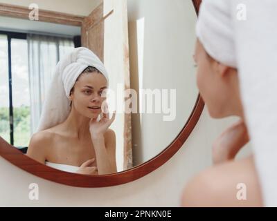 A beauty woman stands in front of a mirror after a shower in a towel on her head looks at her reflection and does a facial massage applies a day cream Stock Photo