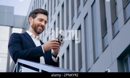 Stylish handsome businessman holding phone reading email stands on street smiling writes message communicates in social network pensive man browses Stock Photo
