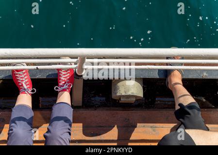 Top view showing legs of two persons, sitting on a narrow sea side passage of a ferry boat, looking toward the sea, and putting their legs on passage handrail Stock Photo