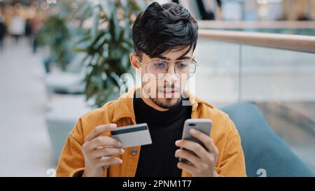 Focused indian man guy customer pay by card on phone using e-banking mobile application arabian male shopper making buying in online store paying Stock Photo