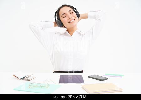 Smiling businesswoman in headphones, sits at office desk listens music and relaxes, rests after work, white background Stock Photo