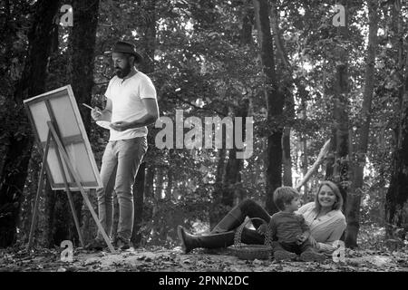 Handsome bearded man draws a picture of his young family in the park using a palette with paints and a spatula. Drawing easel and canvas with a Stock Photo