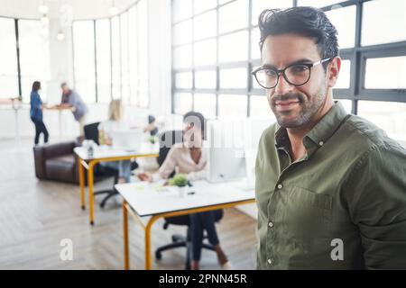 I face challenges head-on. a male designer in a office with his colleagues in the background. Stock Photo