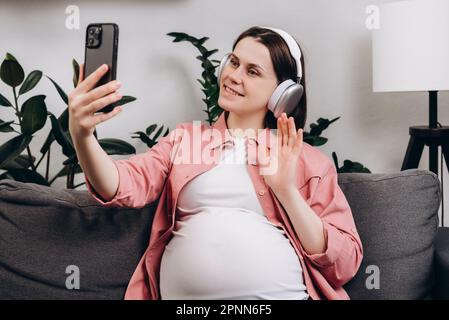 Cheerful cute young pregnant woman with headphones using mobile phone, making video call to friends or relatives, chatting online, future mother wavin Stock Photo