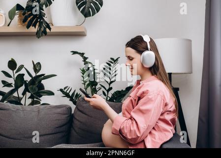 Smiling beautiful young pregnant woman listening to music through wireless headphones, happy future mother using smartphone while resting on sofa in l Stock Photo