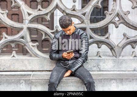 Young man, dressed all in black, consulting his smartphone, seated at the foot of the balustrade of the King's House on the Grand Place in Brussels. Stock Photo