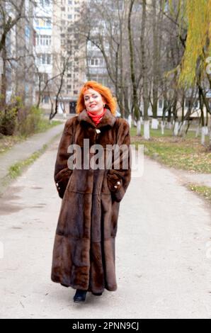 red-haired woman wearing white knitted hat and dressed in warm hooded casual parka jacket outerwear, girl in the parking lot, pedestrian crossing and car in the background, girl warms frozen Stock Photo