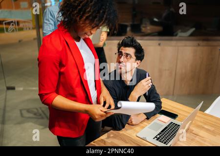 High-angle view of African American female intern, assistant or employee handing out information document to male team leader during boardroom meeting Stock Photo