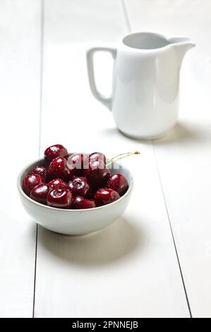 Sweet cherries in a ceramic bowl on wooden background, closeup. Fresh ripe sweet cherries in a bowl with droplets of water and pitcher Stock Photo