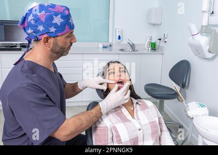 Side view of concentrated ethnic adult male dentist in medical uniform and gloves trying invisible aligners on teeth of female patient sitting on chai Stock Photo