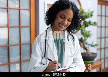 Happy biracial female doctor holding clipboard and taking notes at doctor's office Stock Photo