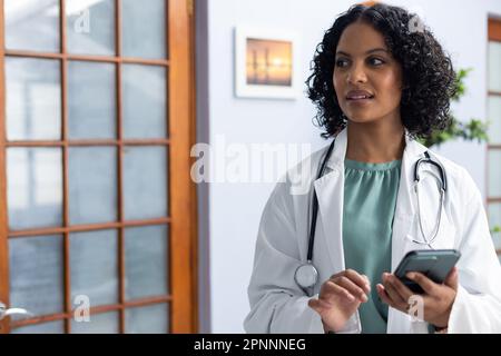 Biracial female doctor wearing sthethoscope, using smartphone at doctor's office Stock Photo