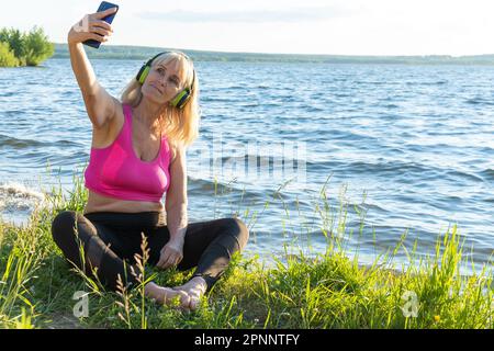 An elderly woman in sports clothes and headphones holds a phone in her hands, watches online exercises for training, waves a greeting with her hand, l Stock Photo