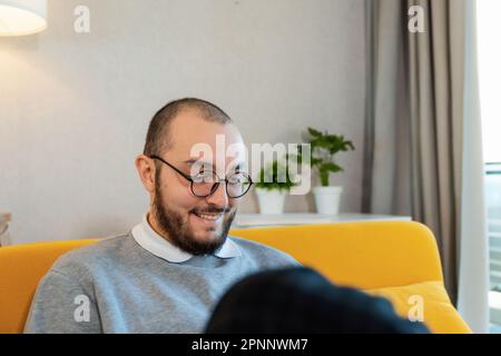 Young attractive hipster man in glasses using mobile phone while sitting on the couch at home. Student using smartphone gadget social media mobile app Stock Photo