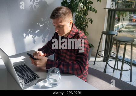 Young caucasian man, digital nomad, freelancer working remotely in cafe, using laptop and smartphone. Male text a message on mobile phone Stock Photo