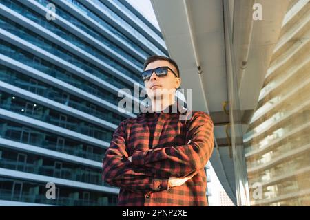 Portrait of positive cheerful reliable freelancer or IT specialist with crossed arms. Man in sunglasses standing outside modern office buildings with Stock Photo