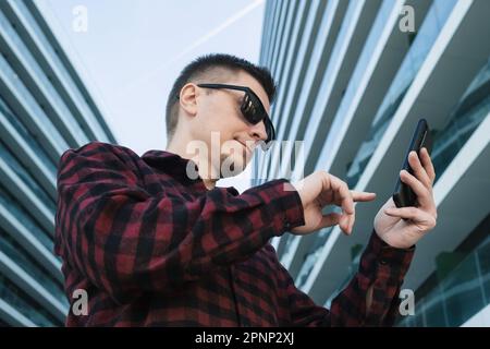 Young man freelancer or IT specialist in sunglasses using mobile phone or smartphone device outside modern office buildings. Hipster guy with a phone Stock Photo