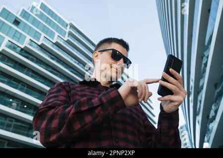 Young man freelancer or IT specialist in sunglasses using mobile phone or smartphone device outside modern office buildings Stock Photo