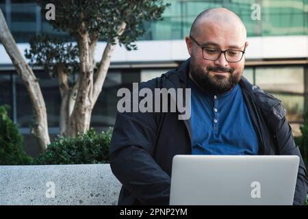 Plus size man in glasses using laptop while working outdoors. Male freelancer or IT specialist using computer for remote work Stock Photo