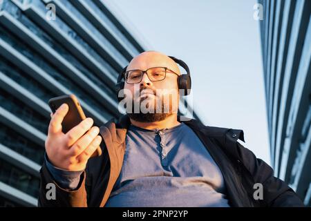 Bearded plus size man in glasses listening music with headphones using phone sitting outdoors with modern office building on background Stock Photo