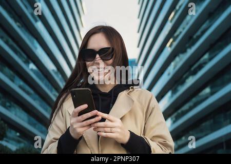 Lifestyle portrait business woman in sunglasses after work and using mobile phone, smiling and typing text message on smartphone device outside the of Stock Photo