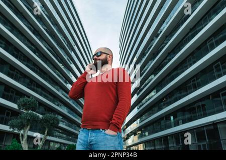 IT guy or freelancer using mobile phone. Bearded man in sunglasses talking on mobile phone outdoor the modern office buildings. Stock Photo