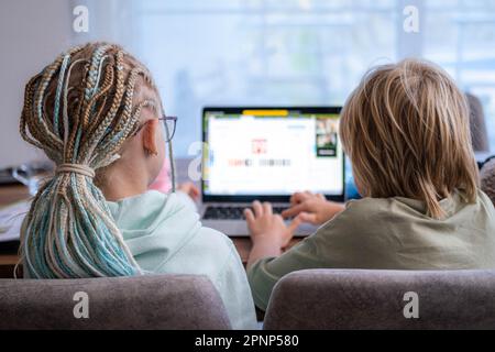 back view siblings pupils boy and girl learning together remotely online at home, looking to laptop, speaking with teacher or tutor using internet Stock Photo