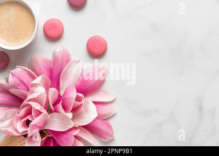 Holiday or wedding background. Pink magnolia flowers in ice cream cone, coffee cup and macarons on white table. Flat lay. Top view Stock Photo