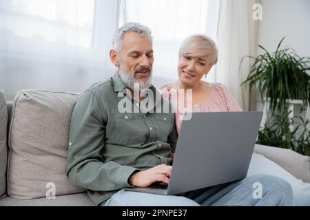 positive mature couple watching movie on laptop while sitting on couch in living room,stock image Stock Photo