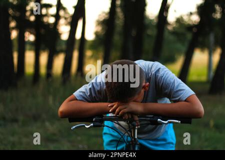 Sad little boy sitting on bicycle near forest. Child cry and hiding his face. Kid alone. Victim of bulling. Problem in school. Social protection. Teen Stock Photo