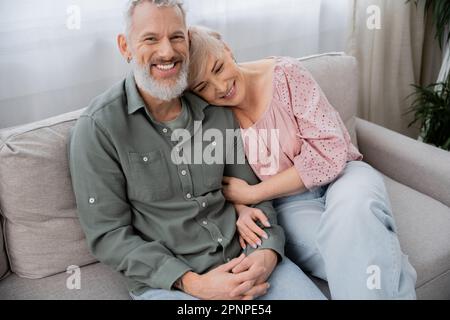 happy middle aged woman leaning on cheerful bearded man smiling at camera on couch in living room,stock image Stock Photo