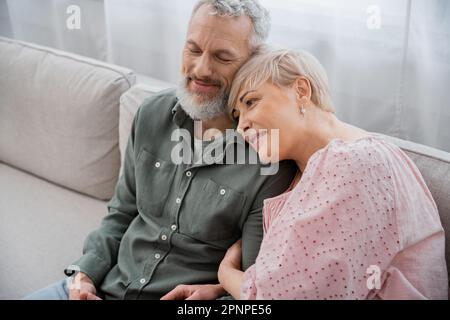 mature woman leaning on pleased bearded husband smiling with closed eyes on couch in living room,stock image Stock Photo