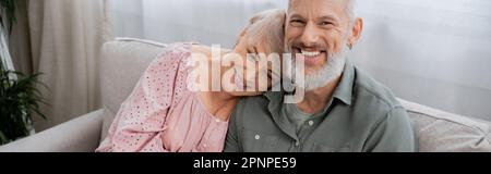 carefree middle aged man smiling at camera near happy wife on sofa in living room, banner,stock image Stock Photo