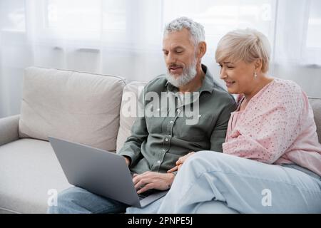 joyful middle aged woman and bearded man sitting on sofa in living room and watching movie on laptop,stock image Stock Photo