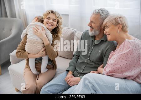 overjoyed woman with closed eyes embracing toddler daughter while spending time with parents on couch at home,stock image Stock Photo