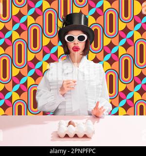 Stylish woman in white blouse, cylinder hat and sunglasses holding candy near lips, eggs on table. Colorful pattern background Stock Photo