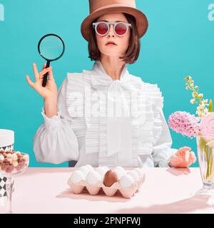 Woman in cylinder hat and sunglasses holding magnifying glass over blue background. Eggs and sweets on table. Holidays preparation Stock Photo