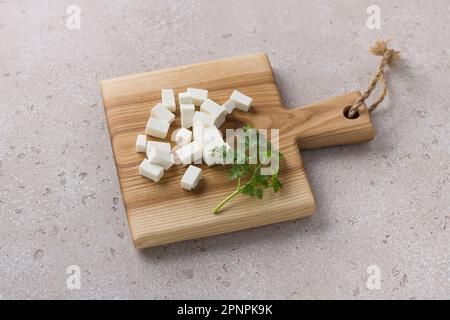 Wooden board with diced feta cheese and a sprig of parsley on a beige textured background. Cooking homemade food Stock Photo