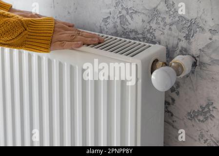 Man in yellow sweater warming his hands on the heater at home during cold winter days. Male getting warm up his arms over radiator. Concept of heating Stock Photo