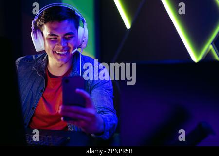 Happy young gamer watching and streaming live game broadcasts on his smartphone. Wearing his headset, he connects and participates in virtual gaming c Stock Photo